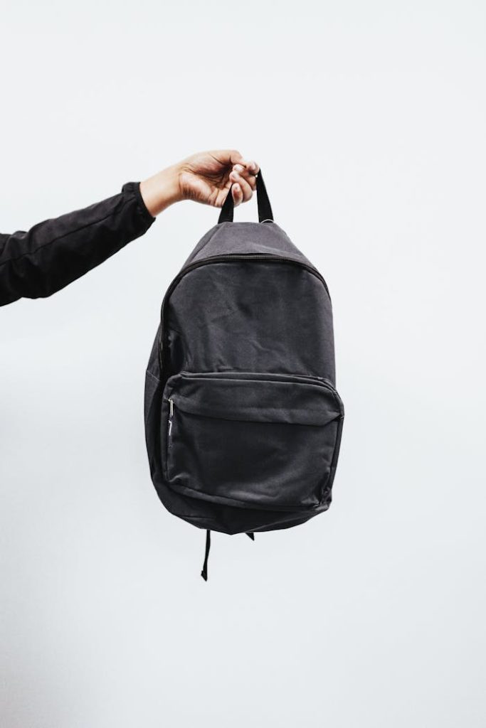 A simple black backpack being held by a hand against a minimal white background, emphasizing style and practicality.